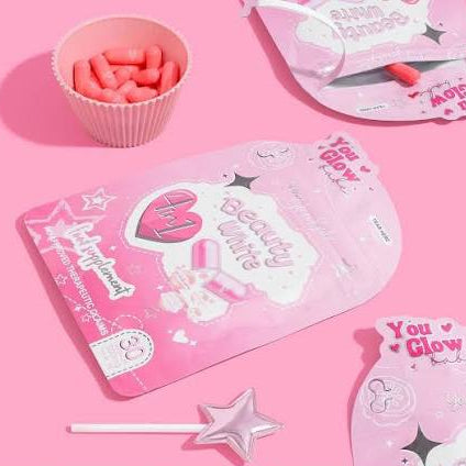 Flat lay aesthetic shot of pink You Glow Babe Beauty White supplement capsules in a cup next to the matching brand packaging on a pink background.