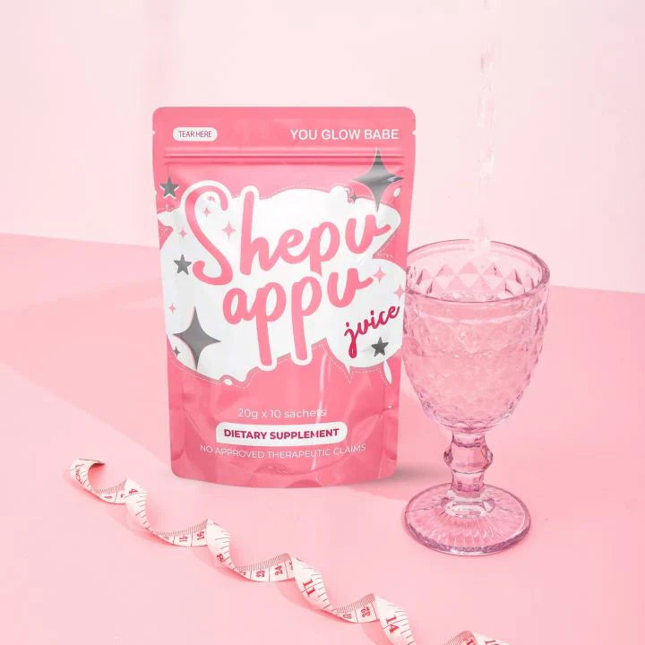 Premium packaging pouch of YouGlow Babe Shepu Appu dietary supplement drink next to an elegant pink crystal glass on a pastel pink background.